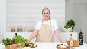 A mature woman in an apron stands at a kitchen counter with fresh produce and cheese, ready for cooking.